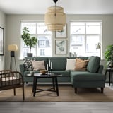 Modern living room with green sofa, rug, wood tables, wicker lamp, plants, and artwork.