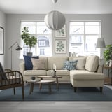 Modern living room with EKHOLMA sofa, armchair, coffee tables, blue rug, and hanging lamp. Large windows, plants, and wall art. Sofa as focal point.