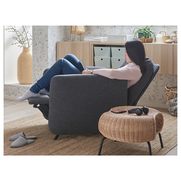 Relaxing in a grey EKOLSUND recliner, feet up on a wicker stool, surrounded by home decor.