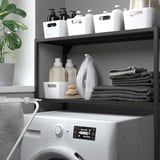 Laundry room with white washing machine under organized shelves, cleaning supplies, folded towels, and a plant.