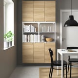 ENHET storage with light wood, white shelf, and decor. Black dining set, plant.