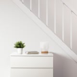Minimalist white dresser against a staircase. Topped with a FEJKA artificial plant, stacked books, and a table lamp.