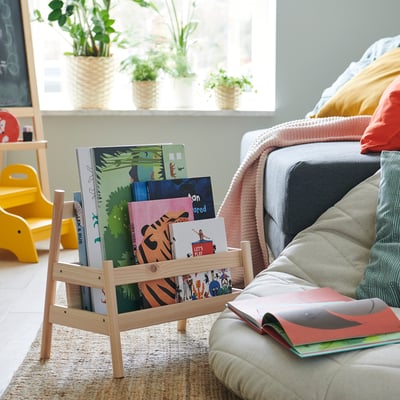 Bright living room with kids wooden bookrack, colorful books, grey couch, yellow chair, plants, and woven rug.