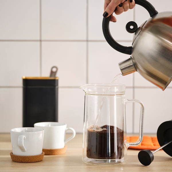 Hand pouring hot water from metal kettle into clear glass coffee maker. Two white ceramic mugs with wooden handles are placed on coasters nearby.