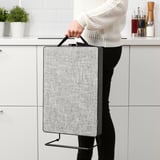 Person holding portable air purifier with handle in kitchen. The purifier has a gray fabric cover and black frame.