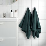 FREDRIKSJÖN dark green towels hang neatly near sink, showcasing their thick, 100% cotton texture and elegant woven border, larger than nearby items.