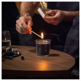 A person lights a small glass candle on a dark wooden table. The candle emits a soft light. A match and various small decorative items are around.