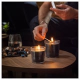 Two hands light scented candles on a wooden table, creating a cozy ambiance with woodsy streams scent. Candles are in clear glass containers.