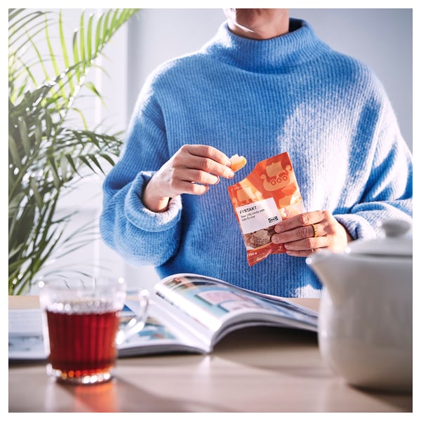 Person in blue sweater holding FYRTAKT candy, sitting at table with tea, plant, and magazine.