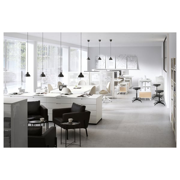 Modern office space with white desks, black chairs, and hanging lamps. Large windows provide natural light.