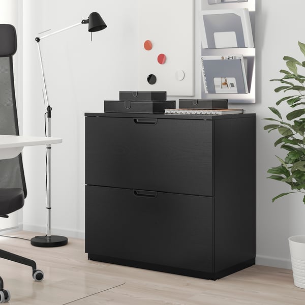 Black GALANT file cabinet with 2 drawers beside white desk. Trays on top hold office supplies. Black lamp and chair complete minimalist setup.