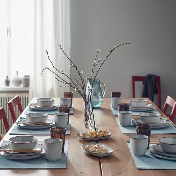 Dining table set with GLADELIG sandy bowls, cups, and saucers. Light blue place mats, red cups, and central vase with branches.