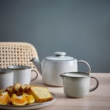 A white tea set with a teapot, two cups, and a cream jug on a table, accompanied by an orange and cake slices on a plate.