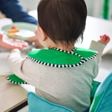 Child wearing a green, waterproof bib with checkered trim sits in a high chair, eating. The bib is adjustable and easy to clean.