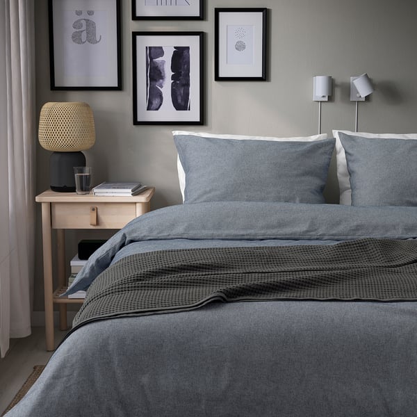 Grey duvet set on bed with pillows, blanket, nightstand with lamp, water, books, art above, wall lights.