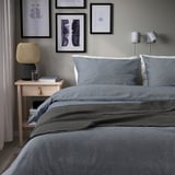 Grey duvet set on bed with pillows, blanket, nightstand with lamp, water, books, art above, wall lights.