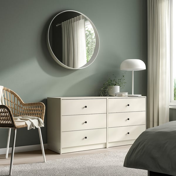 Bedroom with white GURSKEN chest of 3 drawers beside bed, topped with lamp and plant, wicker chair, round mirror.