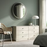 Bedroom with white GURSKEN chest of 3 drawers beside bed, topped with lamp and plant, wicker chair, round mirror.