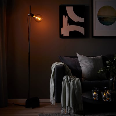 Dark room with HÅRSLINGA black floor lamp, gray couch, blankets, and wall art.