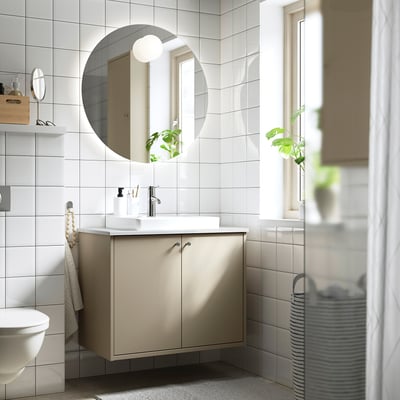 Modern bathroom featuring beige HAVBÄCK vanity, round mirror, white fixtures, greenery, and striped laundry basket.