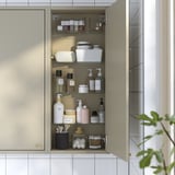 A beige wall cabinet with glass shelves, storing various toiletries like bottles, jars, and brushes. Hinged dampers close the door silently.