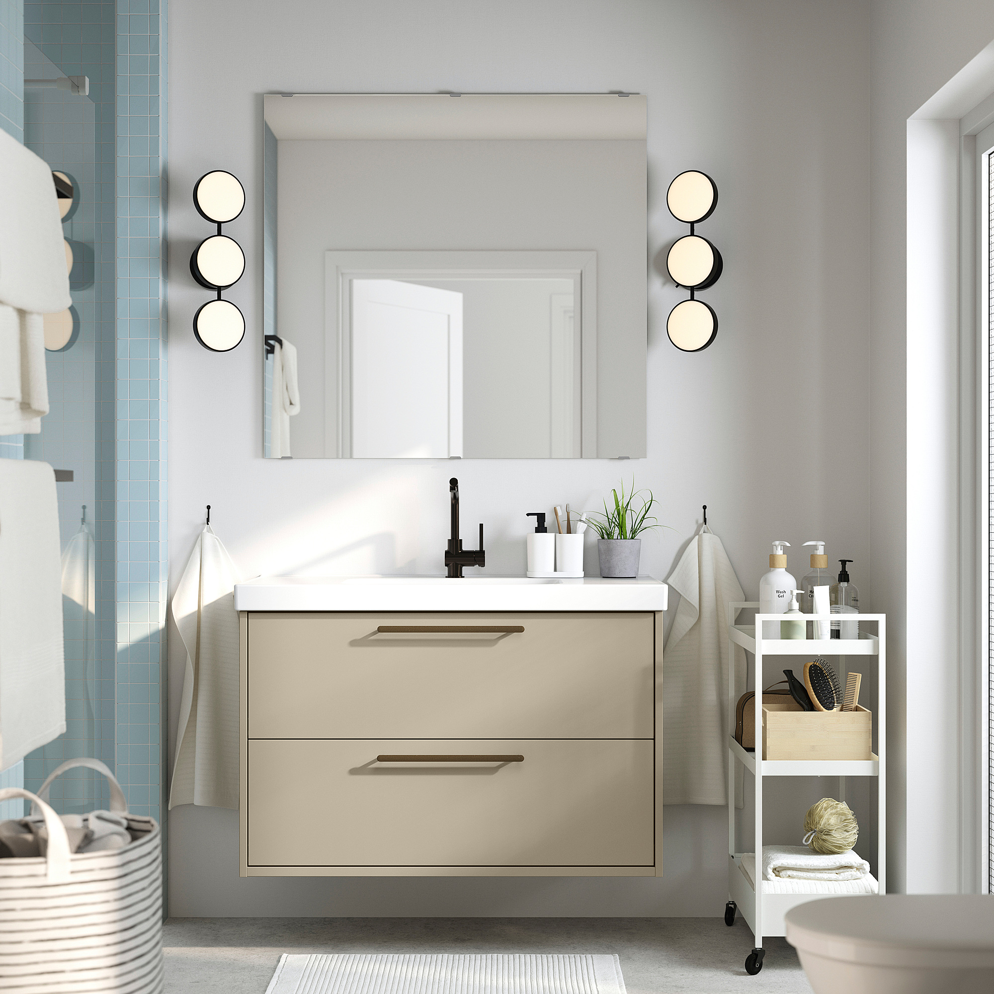 Beige HAVBÄCK vanity, white counter, black faucet, large mirror, two drawers, wall lights, and side cart for toiletries.