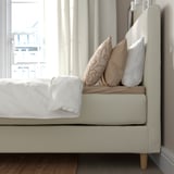 Bed with plush headboard, white pillows, patterned beige pillow, and white blanket, offering comfort and support.