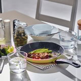 Table setting with HEMLAGAD frying pan filled with vegetables and meat, accompanied by bowls, glasses, and jars.