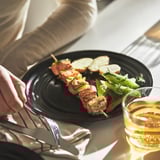 Hand holding plate with meat skewers, vegetables, and bread. Glass of drink beside the plate on a table.
