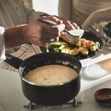 Person serving skewered veggies from black plate beside creamy saucepan on counter.