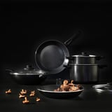 Four modern pans sit together on a dark surface. One wok holds mushrooms, with others stacked or nearby, showing various cooking tools.