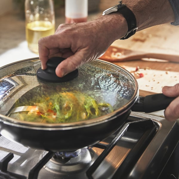 Person lifts lid from sizzling wok on stove, revealing colorful vegetables inside. The glass lid allows monitoring of cooking progress.