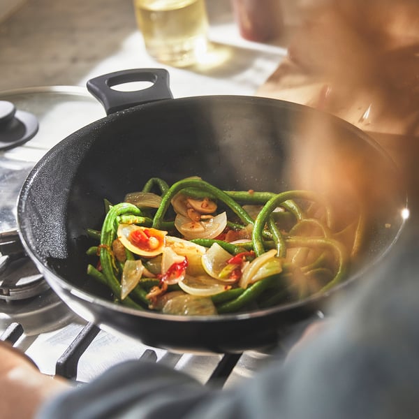 Hand holding wok with stir-fried veggies and mushrooms over gas stove.