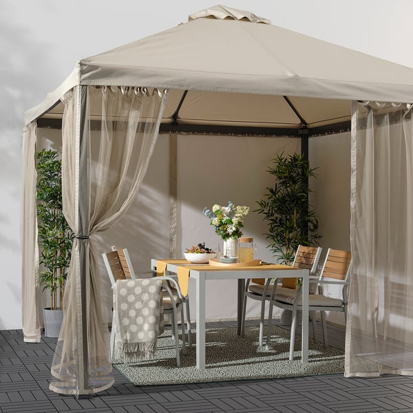 Beige gazebo with sheer polyester curtains, fitting multiple chairs and table, creating cozy ambiance.