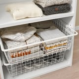 White storage shelf with wire basket holding neatly folded clothes, part of ikeas HJÄLPA organizational system.