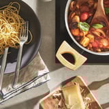 Black pan with spaghetti, fork, wooden handles. Nearby: saucepan with veggies, cheese on wood.