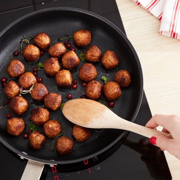 A hand stirs golden-brown plant-based meatballs in a black skillet, garnished with herbs and red berries.