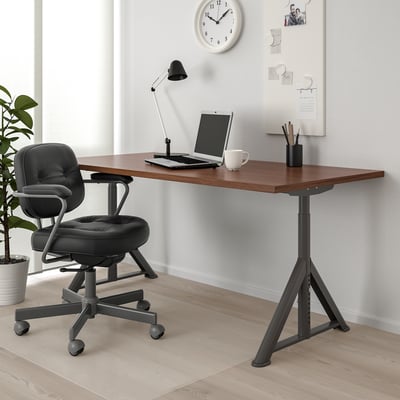 IDÅSEN desk with dark top, black legs. Laptop, mug, lamp on it. Black swivel chair. Wall clock, corkboard.