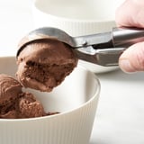 Person scoops chocolate ice cream using an IDEALISK tool into a white bowl.