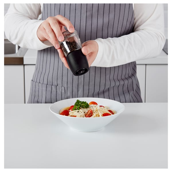 Person grinds pepper with black IKEA mill over pasta bowl.
