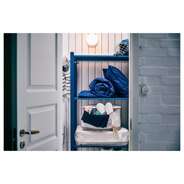 Blue wooden storage unit against a light brick wall, with white and blue bed linens and a lamp on top shelf.