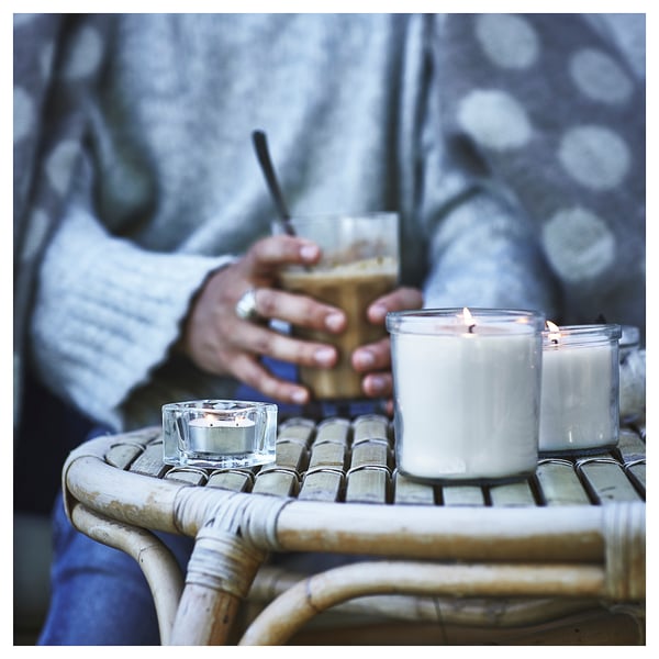 Person holding white candle on wicker table. Two lit candles on table, one in glass. Background has dotted fabric.