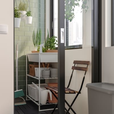 Small balcony with JOSTEIN white metal storage unit holding potted plants and supplies. Wooden folding chair, green tiled wall, and a window.