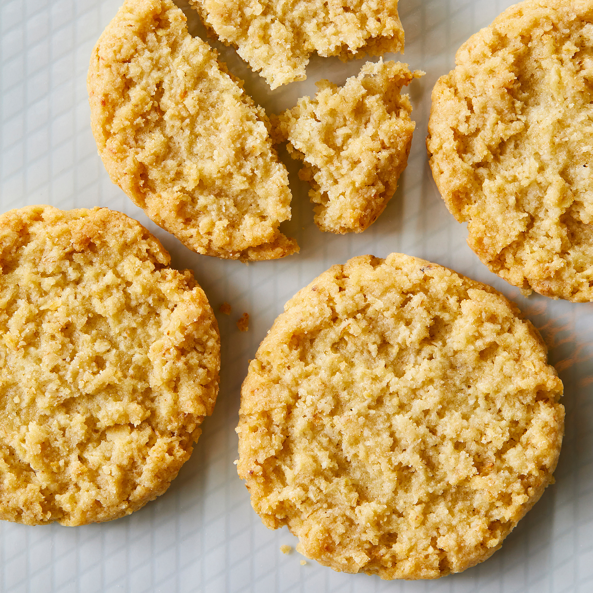 Six round, golden-brown oat biscuits on a white surface. One is broken, showing crumbly texture.