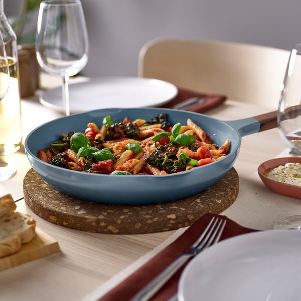 Blue cast aluminum frying pan with wooden handles on a table, surrounded by plates, glasses, and bread. Perfect for serving.