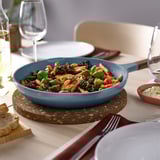 Blue cast aluminum frying pan with wooden handles on a table, surrounded by plates, glasses, and bread. Perfect for serving.
