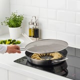 Person holding a splatter screen over a pan with food frying, preventing splashes.