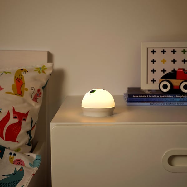 Annimal-patterned pillow sits on a white drawer. A small, round nightlight named KORNSNÖ glows softly beside stacked books.