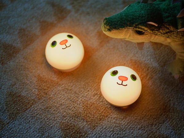 Three round, white night lights with cartoon faces in a soft, warm glow, next to a green dinosaur plush toy on a carpeted floor.