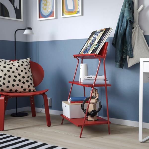 Red KRÖNIKÖR shelf with books, speaker, & chair with pillow against blue wall, coat rack nearby.